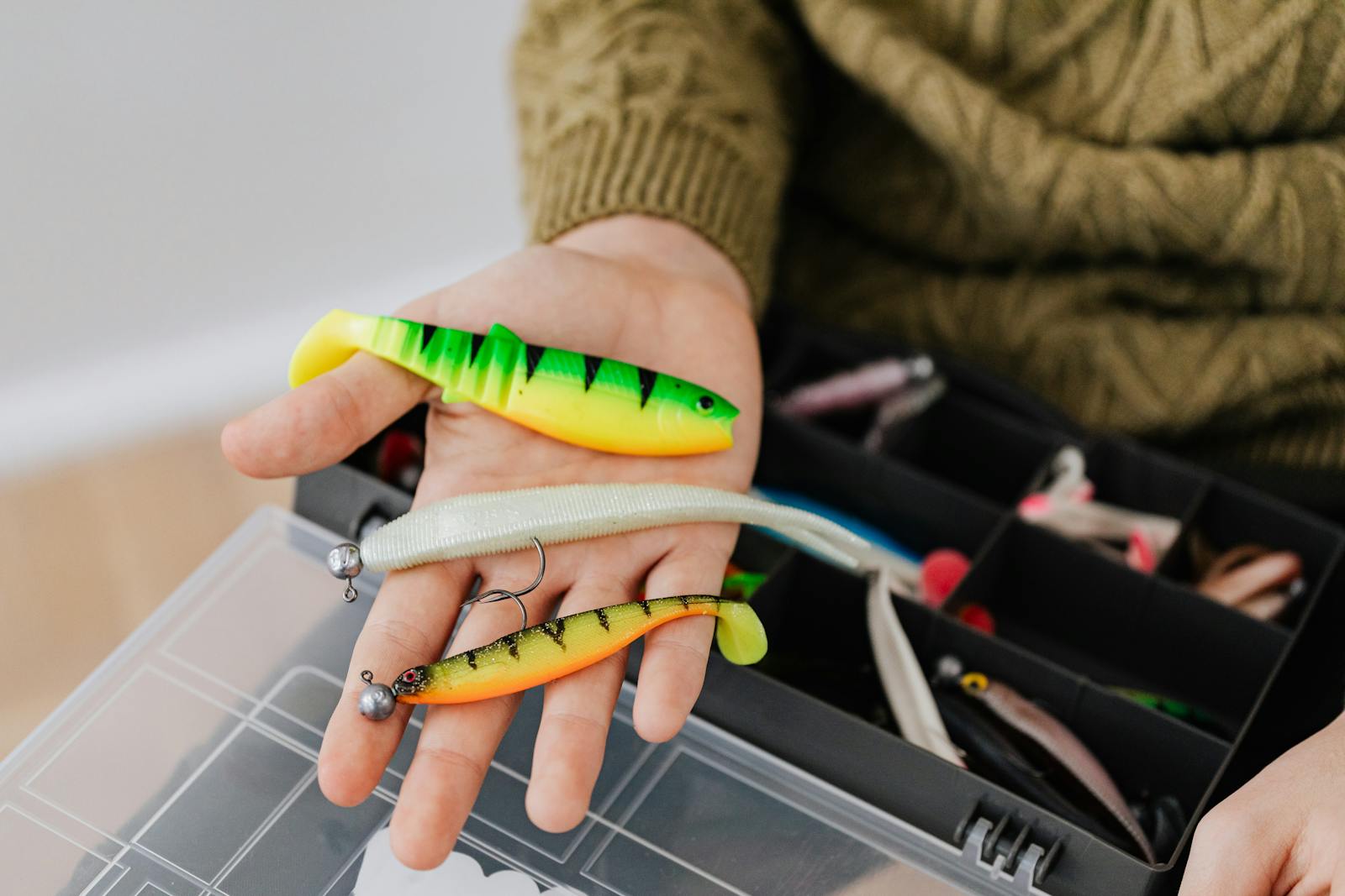 Close-up of colorful fishing lures in a hand, next to a fishing tackle box.