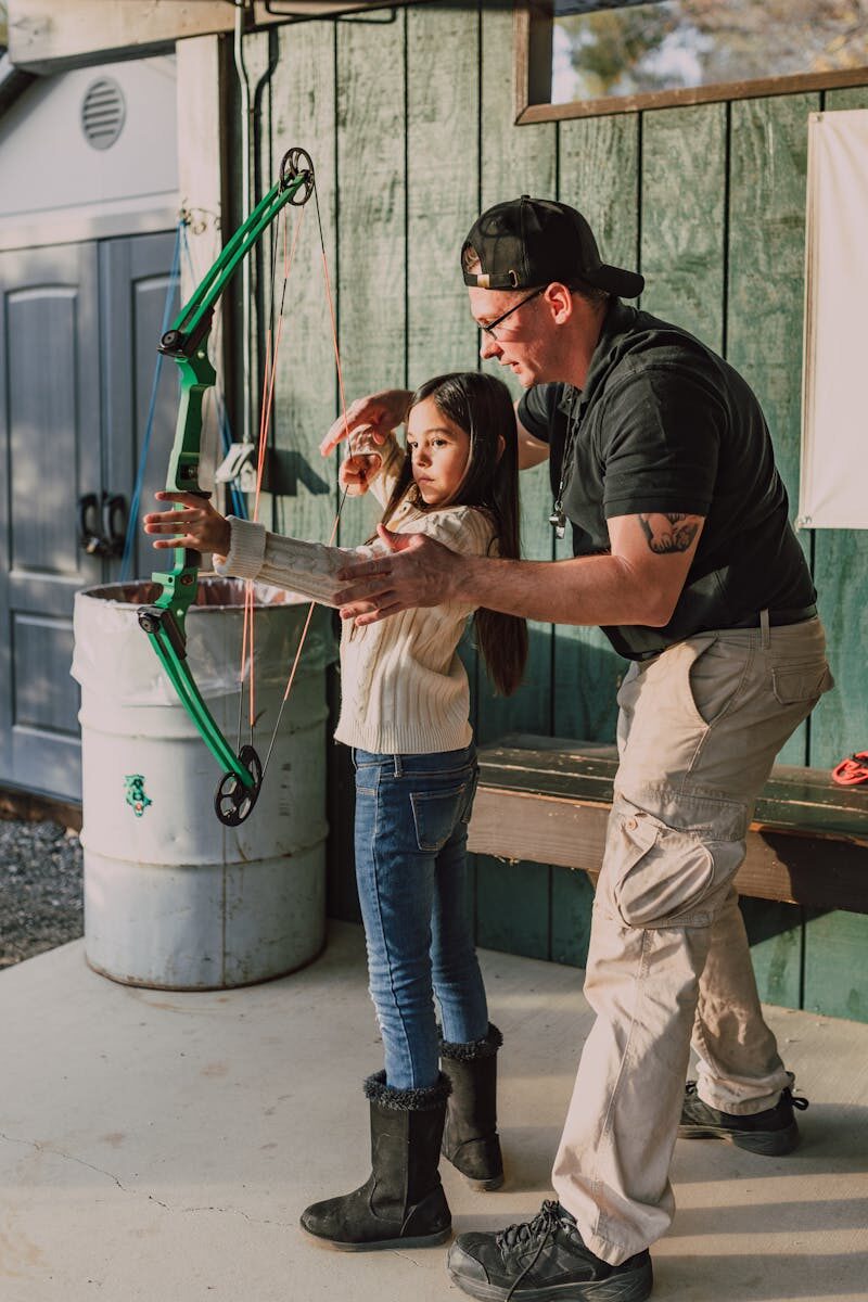 My Neighbor Got Mad Because I’m Teaching My Daughter Archery A young girl practices archery with guidance from an instructor in an outdoor setting.